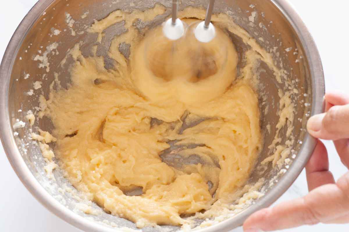 Beating an egg and vanilla into butter and sugar with a hand mixer in a metal bowl.