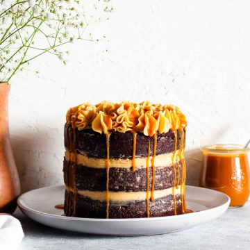 A three layer chocolate salted caramel cake on a white plate next to a vase with white flowers and a jar of caramel.