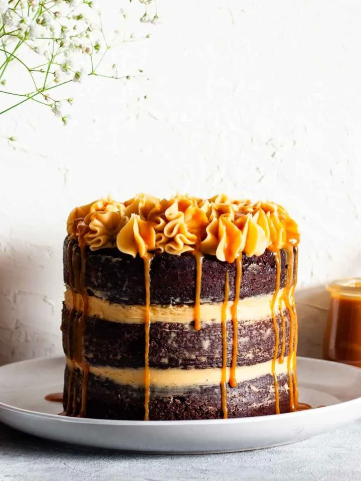 A three layer chocolate salted caramel cake on a white plate next to a vase with white flowers and a jar of caramel.
