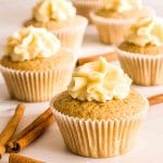 Cinnamon cupcakes with cream cheese frosting on a white surface next to cinnamon sticks.