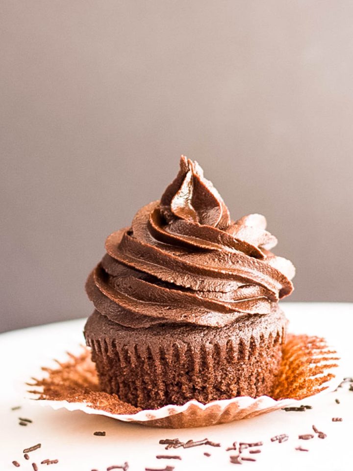 A classic chocolate cupcake with chocolate frosting on a white surface with chocolate sprinkles.