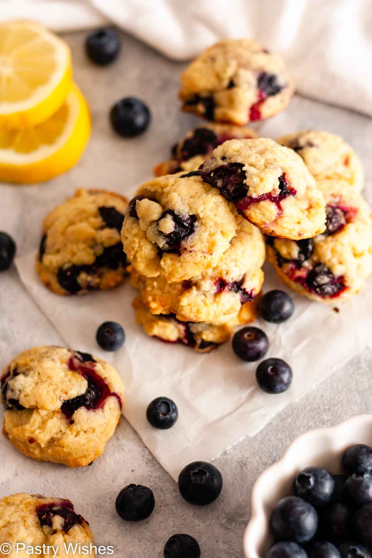 A stack of lemon blueberry cookies on parchment paper next to a bowl of blueberries and sliced lemons.