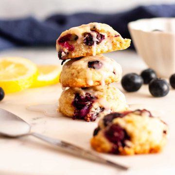 A stack of three lemon blueberry cookies on a white surface dripping with lemon glaze next to a cookie, blueberries and lemon slices.