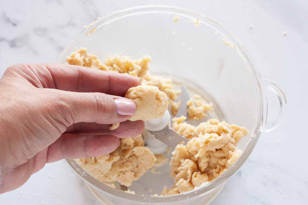 A hand holding a piece of dough and tart dough in a food processor.