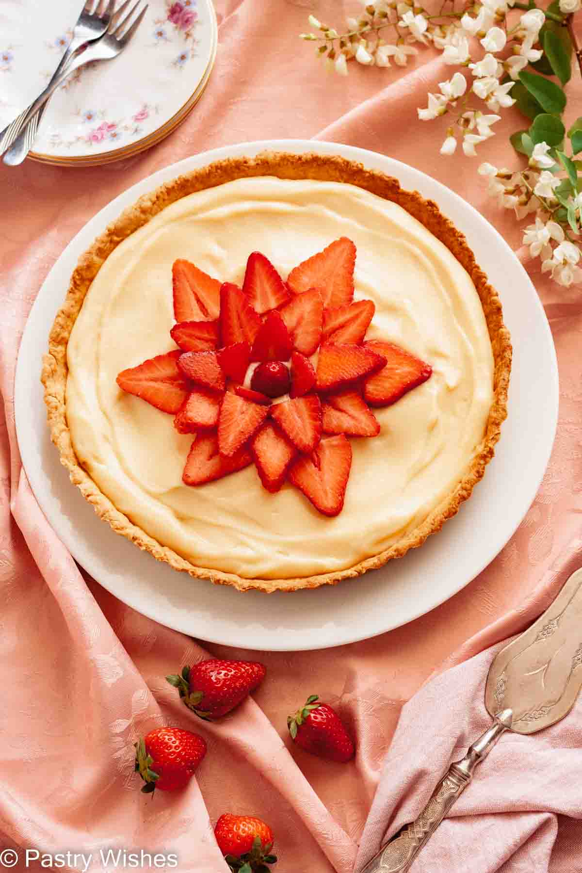 An overhead photo of a tarte aux fraises, French strawberry tart, on a pink tablecloth next to flowers and strawberries.