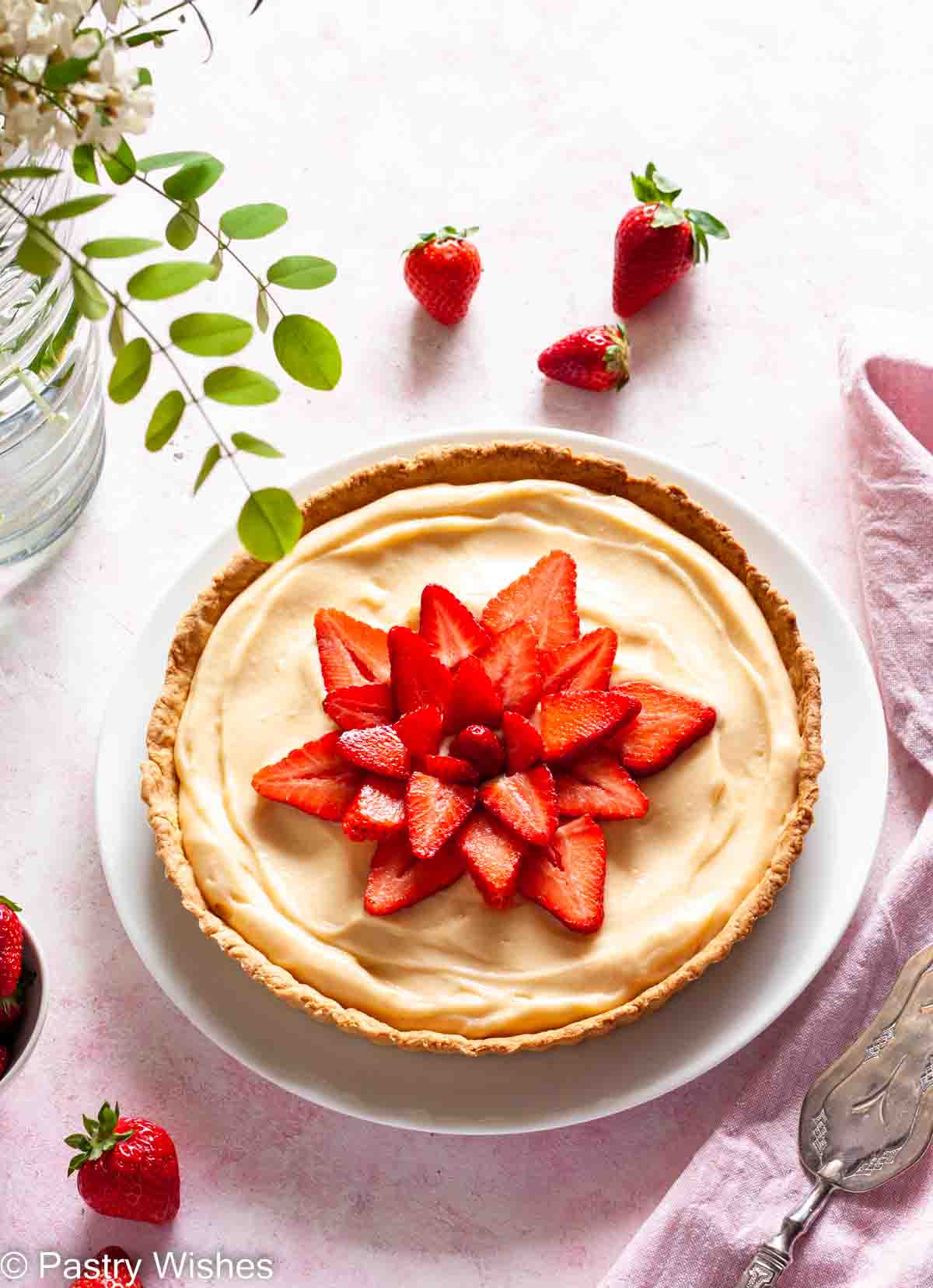 An overhead photo of a tarte aux fraises, French strawberry tart, on a pink surface next to strawberries and a vase filled with flowers.