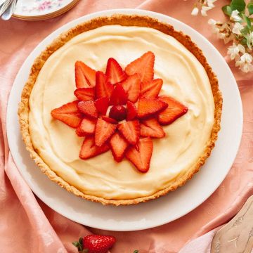 An overhead photo of a tarte aux fraises, French strawberry tart, on a pink tablecloth next to flowers and strawberries.