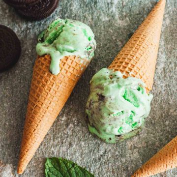 Mint oreo ice cream on two ice cream cones on a gray and white surface next to oreo cookies, mint leaves and an ice cream scoop.