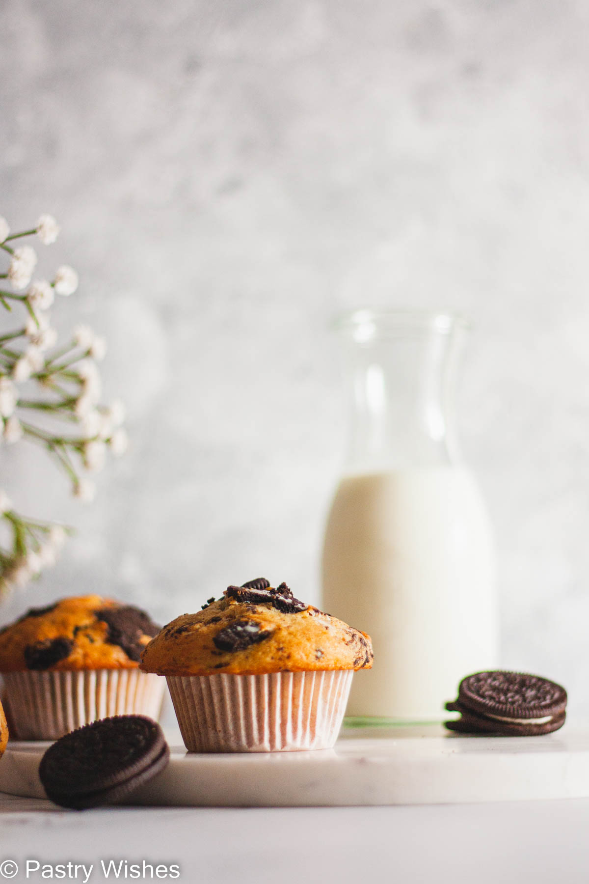 Two Oreo muffins next to white flowers, a bottle of milk and Oreo cookies.
