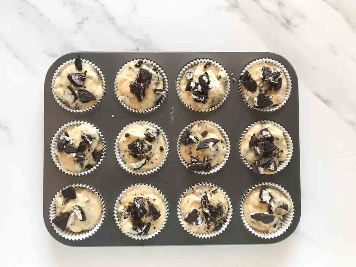 Paper liners in a cupcake pan filled with Oreo muffin batter and topped with crushed Oreo cookies.