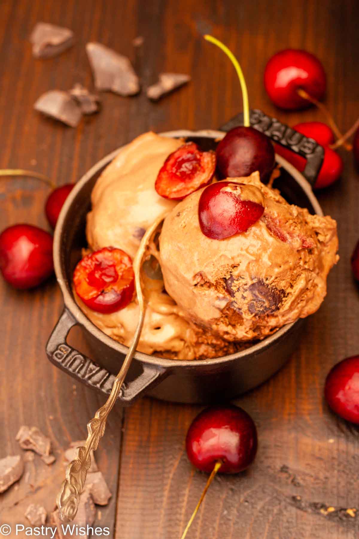 Black forest ice cream topped with cherries in a black bowl on a wooden surface next to cherries and chocolate pieces.