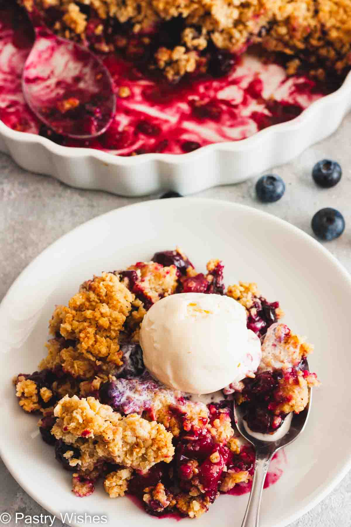Blueberry apple crumble topped with vanilla ice cream with a baking dish filled with crumble behind it.