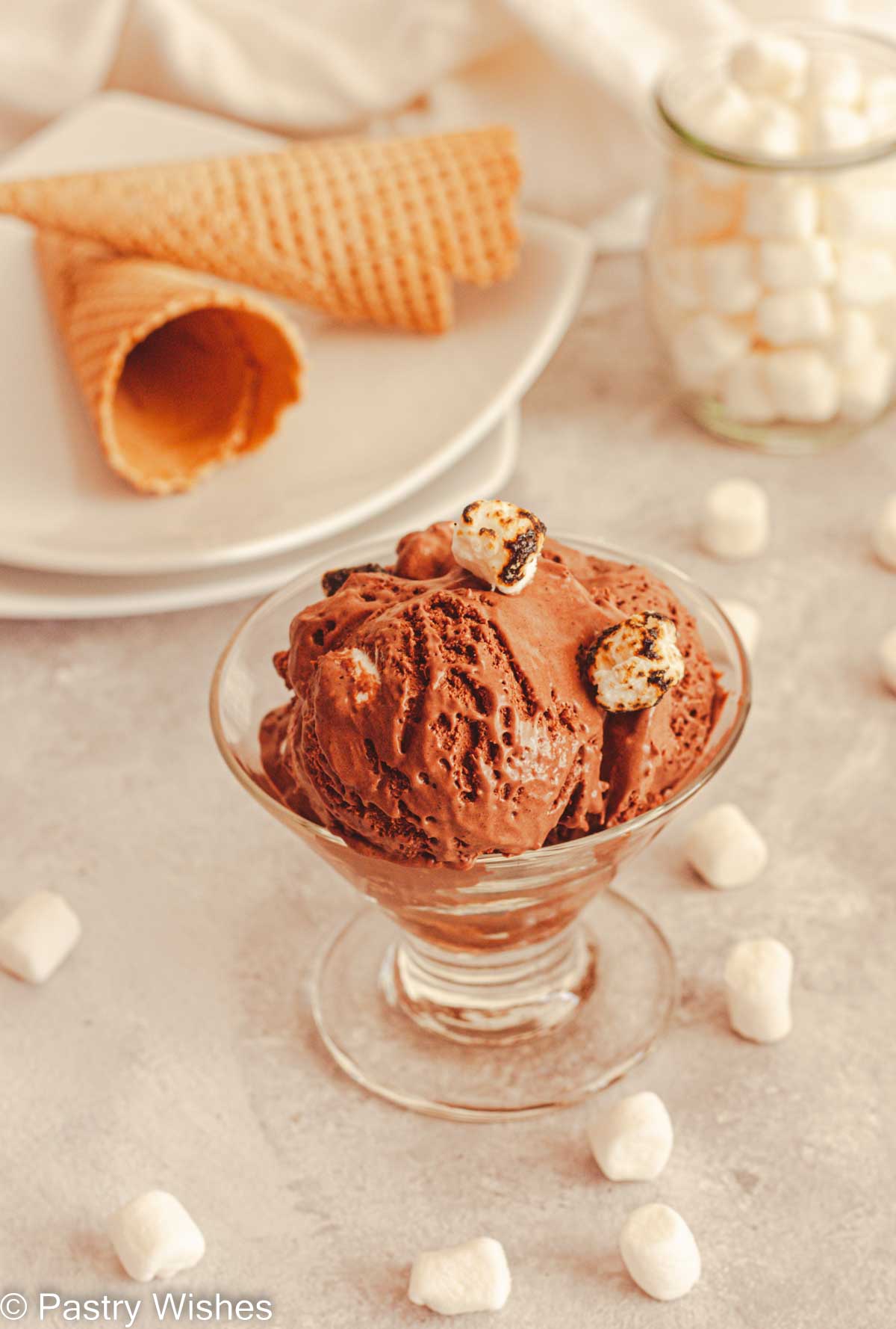 Chocolate marshmallow ice cream in a glass bowl next to white plates with ice cream cones and a jar filled with mini marshmallows.