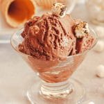 A close up of chocolate marshmallow ice cream in a glass bowl topped with two toasted marshmallows next to marshmallows and ice cream cones.