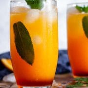 Two glasses of virgin sunrise mocktails with metal straws on a wooden surface, garnished with mint leaves next to a blue towel and orange slices.