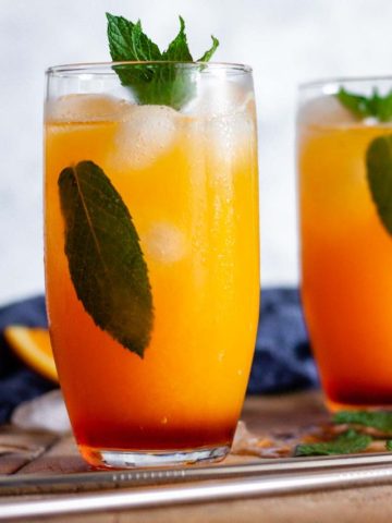Two glasses of virgin sunrise mocktails with metal straws on a wooden surface, garnished with mint leaves next to a blue towel and orange slices.