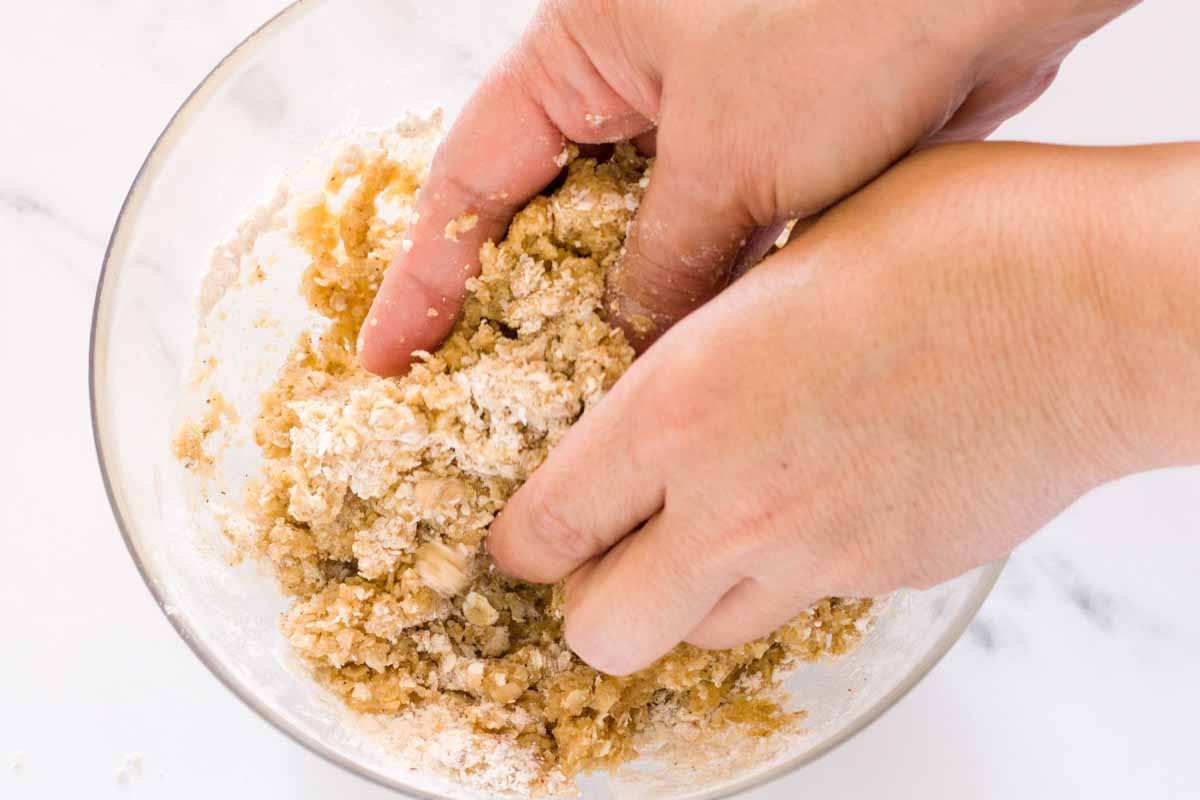 Two hands mixing crumble topping ingredients in a glass bowl.
