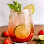 A closeup of a strawberry mojito mocktail on a wooden surface next to strawberries, mint leaves, and lime slices.