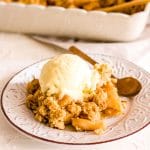 Some vegan apple crisp topped with vanilla ice cream on a brown and white plate with a spoon with a baking dish in background.