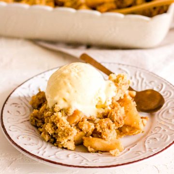 Some vegan apple crisp topped with vanilla ice cream on a brown and white plate with a spoon with a baking dish in background.