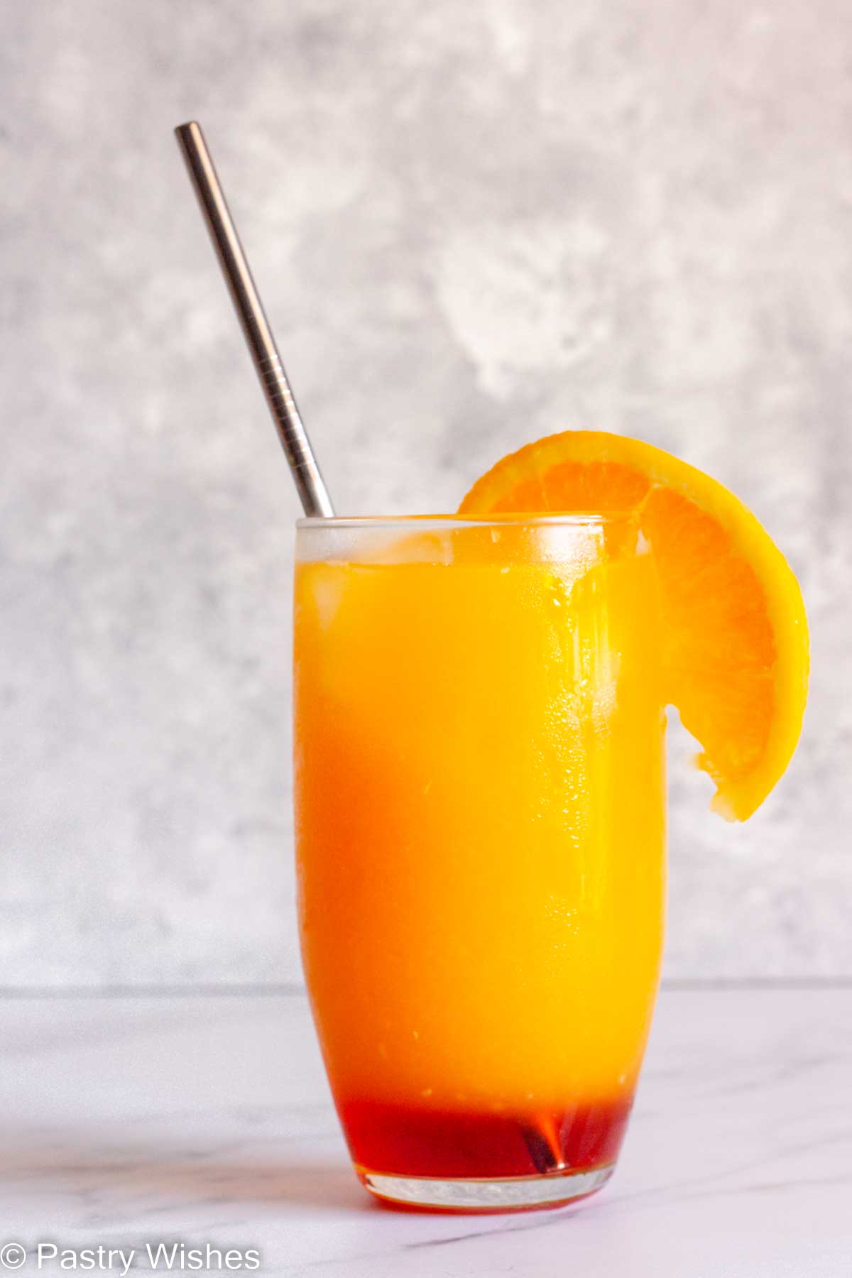 A virgin sunrise in a tall glass with a metal straw garnished with an orange slice.