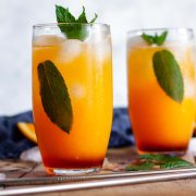 Two glasses of virgin sunrise mocktails with metal straws on a wooden surface, garnished with mint leaves next to a blue towel and orange slices.
