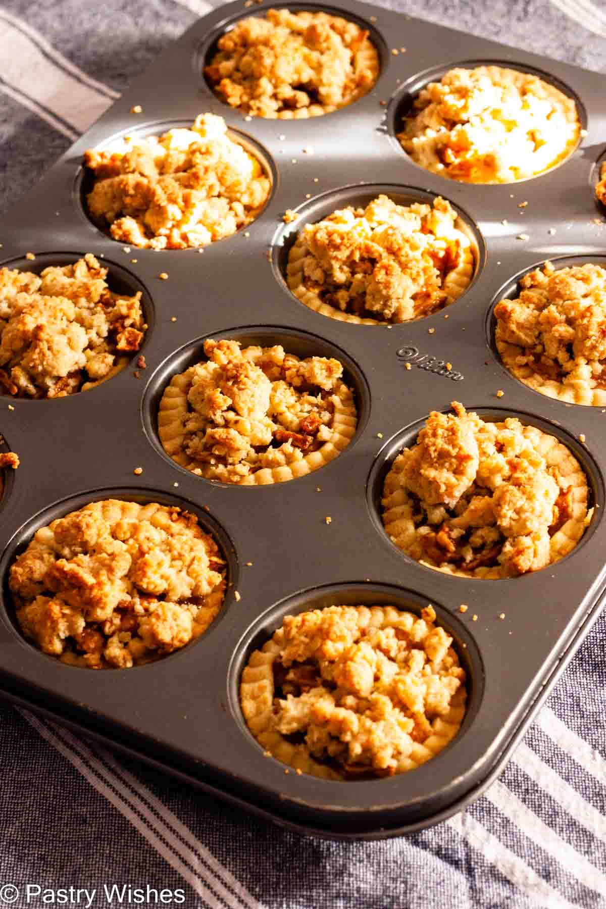 Baked apple pie tartlets in a cupcake pan on a gray and white kitchen towel.