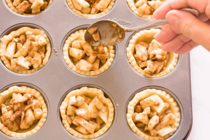 A hand filling mini pie crusts with apple filling using a spoon.