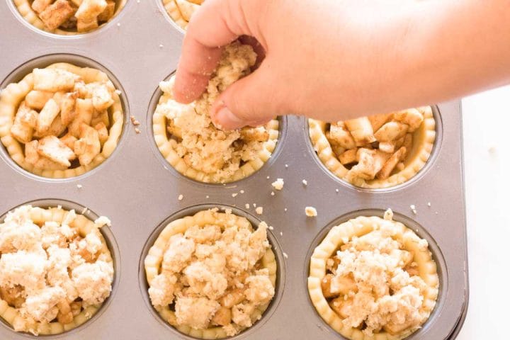 A hand adding crumble topping over mini apple pie tartlets before baking.