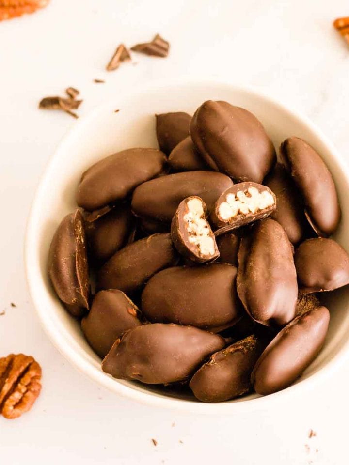 A closeup of chocolate covered pecans in a white bowl with a pecan cut in half.