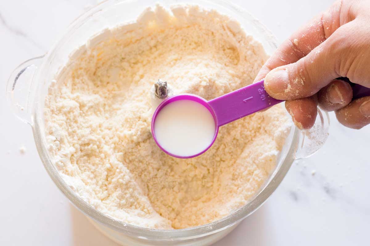 Pouring milk with a measuring spoon over pulsed flour and butter mixture in a food processor.