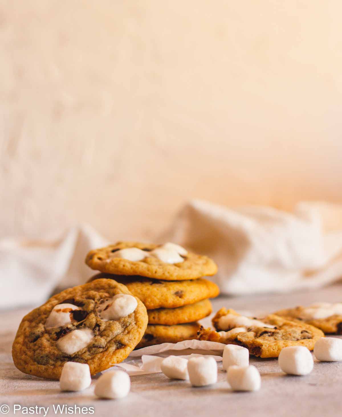 A stack of chocolate chip marshmallow cookies on parchment paper next to mini marshmallows and cookies.