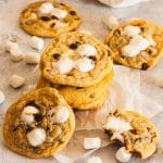 A stack of chocolate chip marshmallow cookies on parchment paper next to mini marshmallows and cookies.