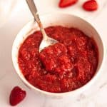 Strawberry filling for cakes in a white bowl with a spoon on a white surface with strawberries.