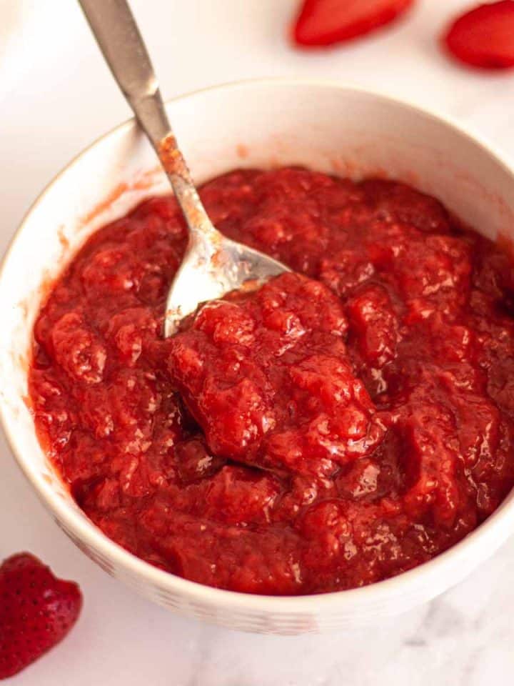 Strawberry filling for cakes in a white bowl with a spoon on a white surface with strawberries.