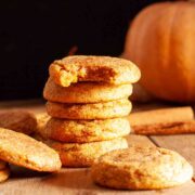A stack of chewy pumpkin cookies on a wooden surface next to a pumpkin and cinnamon sticks and a bitten cookie.