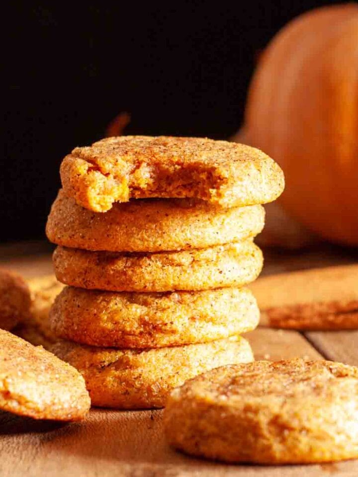A stack of chewy pumpkin cookies on a wooden surface next to a pumpkin and cinnamon sticks and a bitten cookie.
