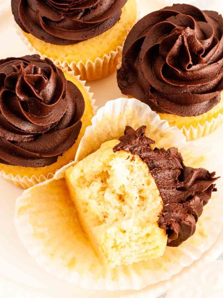 Four vanilla dairy free cupcakes with chocolate frosting on a white plate, one bitten into on an unwrapped paper liner.