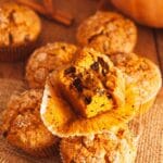 A close up of a bitten pumpkin banana muffin on a paper liner on top of muffins on burlap with a pumpkin and cinnamon sticks in background.