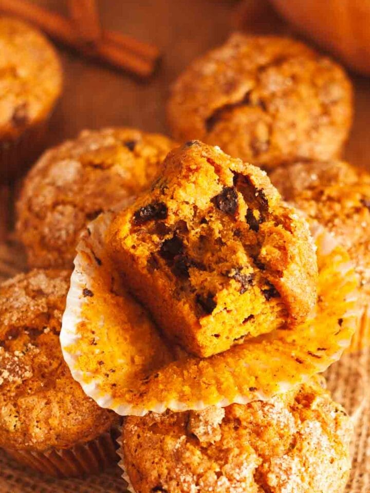 A close up of a bitten pumpkin banana muffin on a paper liner on top of muffins on burlap with a pumpkin and cinnamon sticks in background.