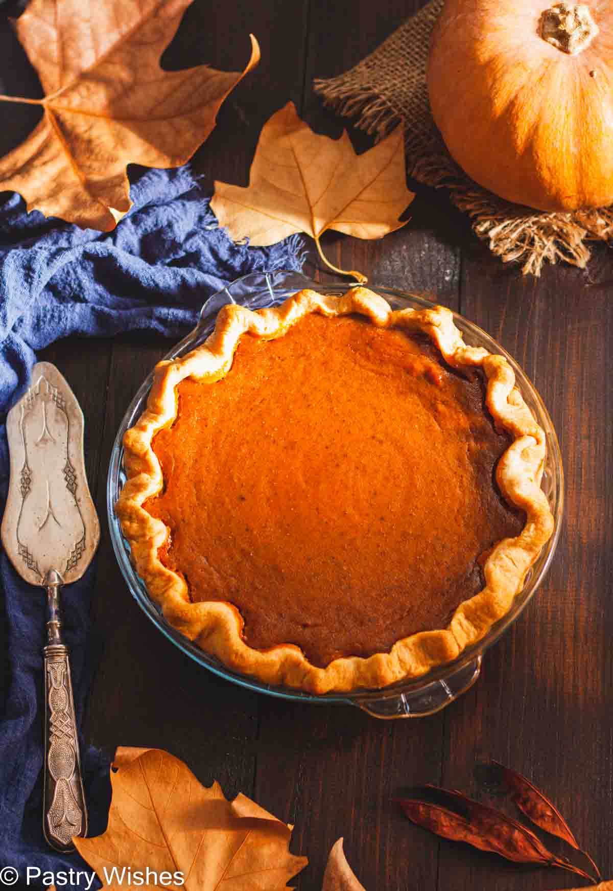 A whole pumpkin pie in a glass pie dish on a wooden surface next to leaves and a pumpkin.