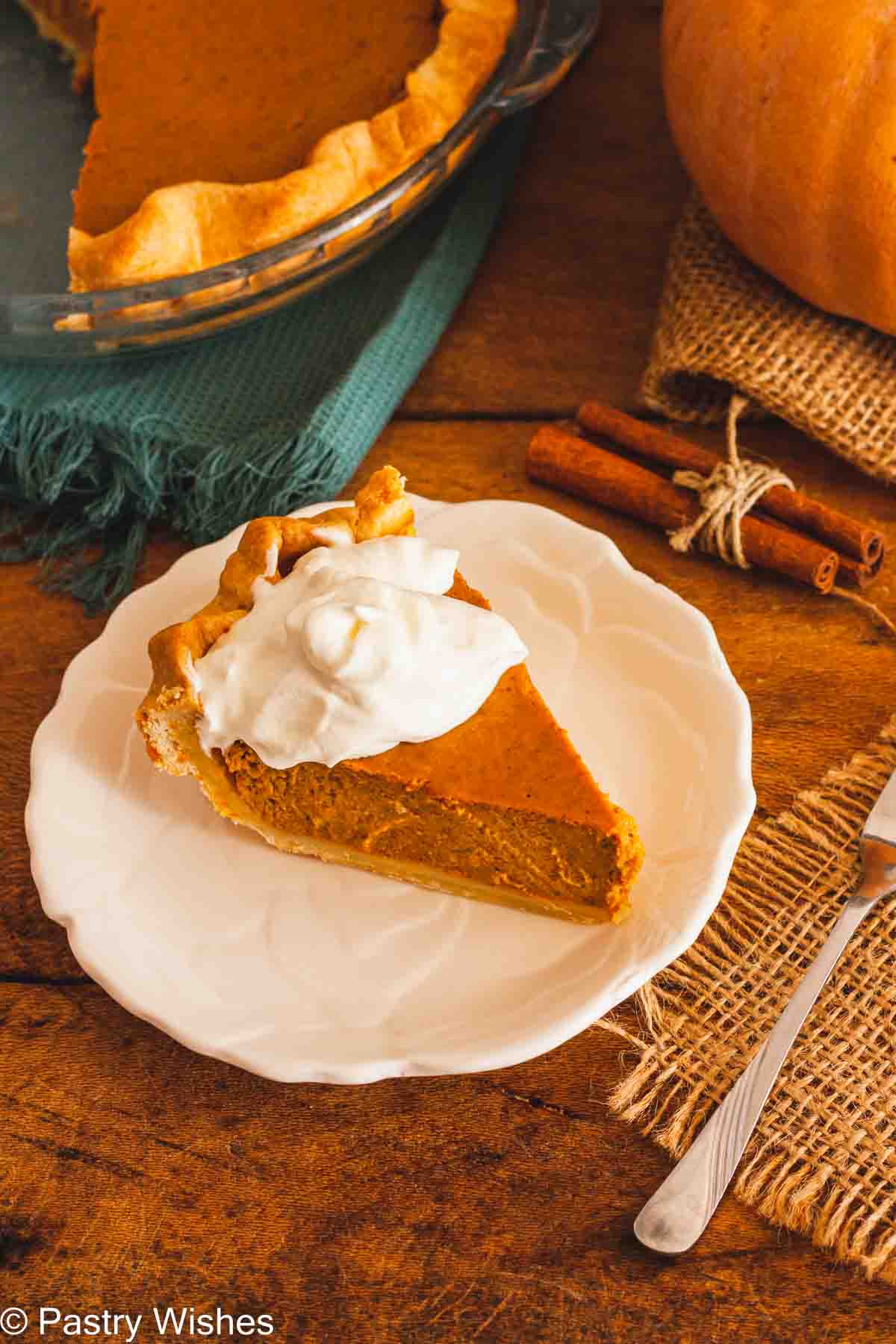 A slice of pumpkin pie on a white plate topped with whipped cream on a wooden surface next to cinnamon sticks and a pumpkin.