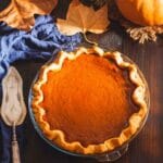 A whole pumpkin pie in a glass pie dish on a wooden surface next to leaves and a pumpkin.