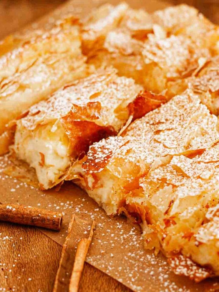 A wooden table with quares of Greek Bougatsa on brown parchment paper dusted with powdered sugar and cinnamon next to cinnamon sticks.