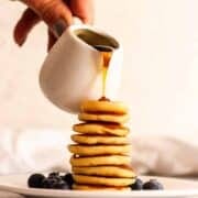 Pouring maple syrup over a stack of mini pancakes on a white plate with blueberries.