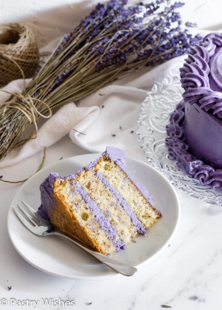 A slice of Earl Grey lavender cake on a white plate next to lavender flowers and cake.