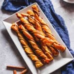 Puff pastry cinnamon twists on a white plate next to a cup of coffee and cinnamon sticks.