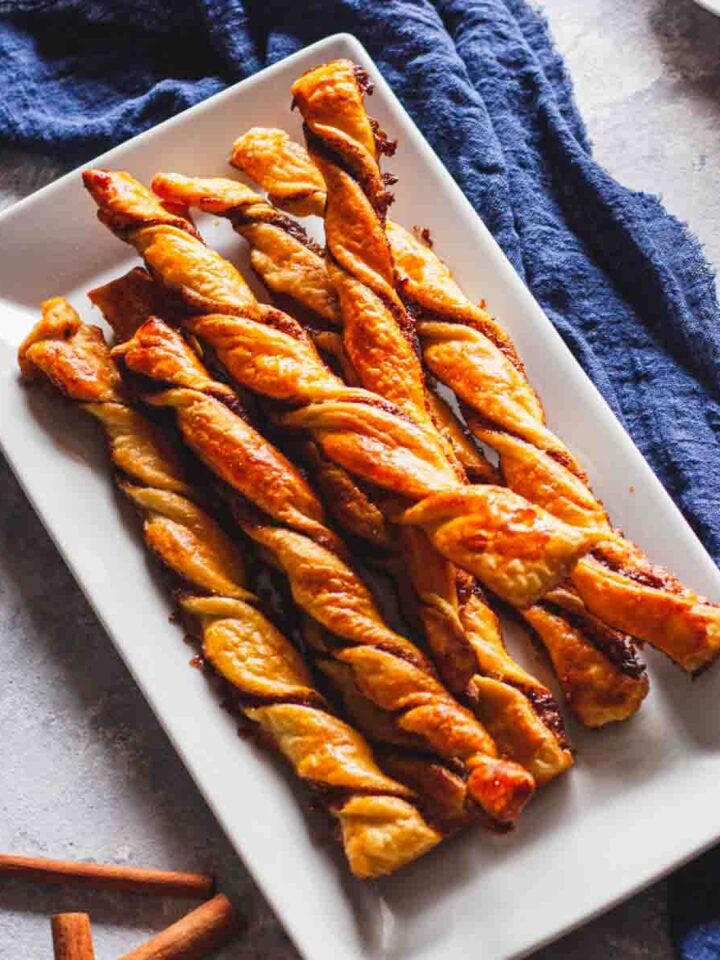Puff pastry cinnamon twists on a white plate next to a cup of coffee and cinnamon sticks.