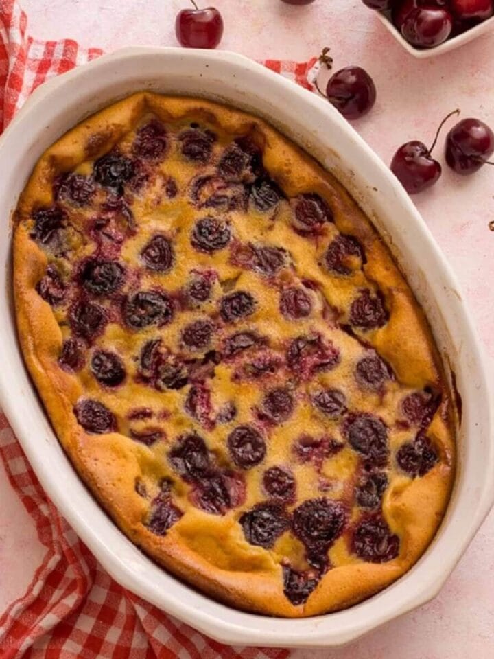 A white baking dish with French cherry clafoutis on a pink surface next to cherries.
