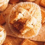 A closeup of a bitten Lotus Biscoff cupcake with Biscoff cookie butter filling dripping in the center on a wooden surface next to cupcakes and cookies.